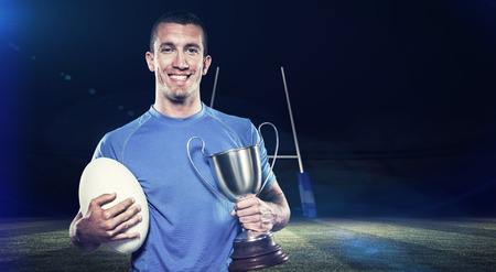 Portrait of smiling rugby player holding trophy and ball against rugby stadiumの写真素材