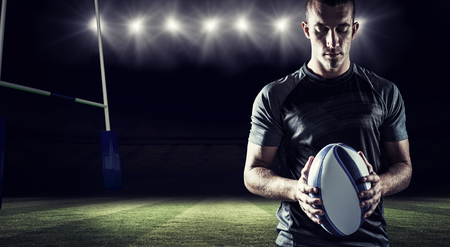 Thoughtful rugby player holding ball against rugby stadiumの写真素材