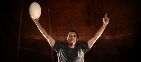Portrait of rugby player in blue jersey holding ball with arms raised against rugby stadiumの写真素材
