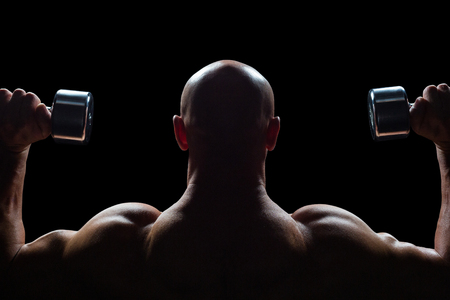 Rear view of man exercising with dumbbells against black backgroundの写真素材