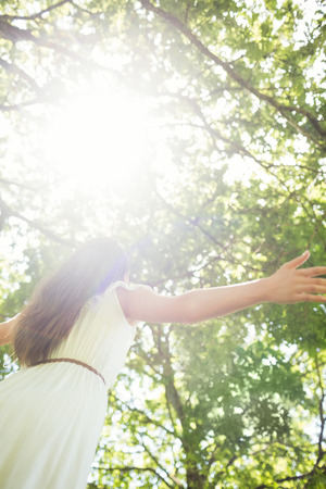 Low angle view of woman with arms outstretched against bright sunlight in parkの写真素材