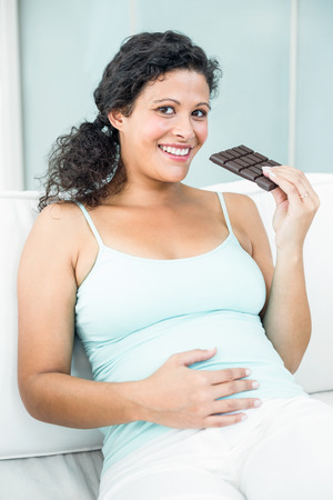 Portrait of happy pregnant woman with chocolate bar sitting on sofaの写真素材
