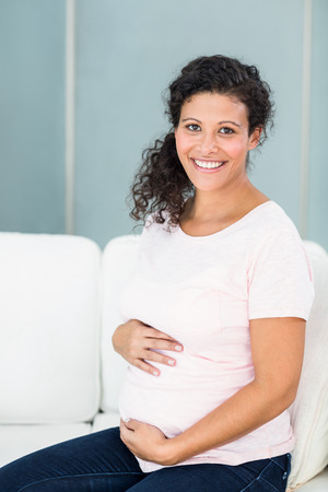 Portrait of happy pregnant woman sitting on sofa against wallの写真素材