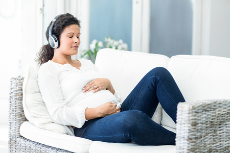 Happy pregnant woman listening to music with closed eyes reclining on sofaの写真素材