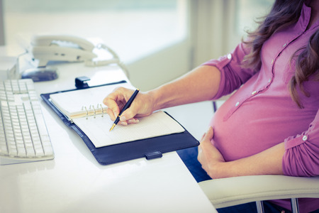 Midsection of pregnant businesswoman writing on diary at desk in home officeの写真素材