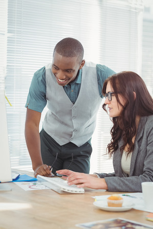Smiling businessman looking at computer while discussing with coworker at desk in officeの写真素材