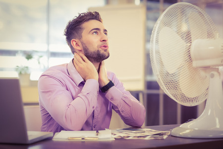 Businessman looking up while sitting at desk with electric fan in officeの写真素材