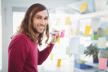 Portrait of hipster writing on sticky note stuck on glass in officeの写真素材