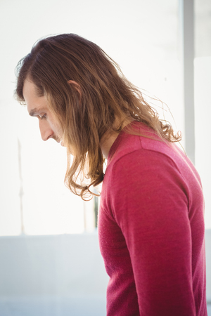 Side view of man looking down while standing against window in officeの写真素材