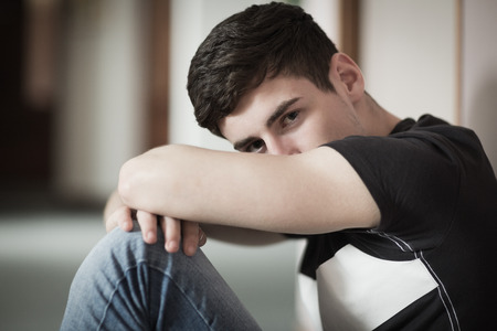Close-up portrait of male student with head on knees sitting by wall in collegeの写真素材