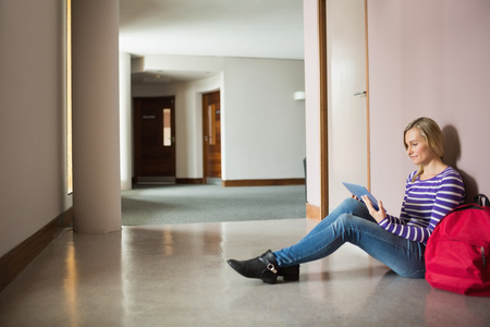 Female student using digital tablet sitting by wall in corridor at collegeの写真素材