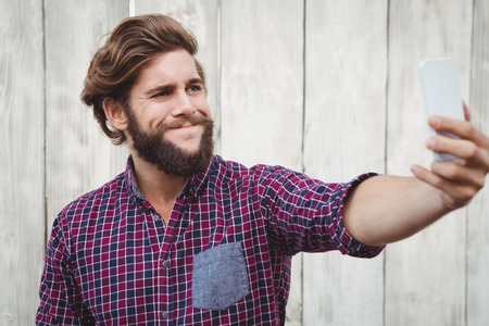 Hipster making face while taking selfie against wooden fenceの写真素材