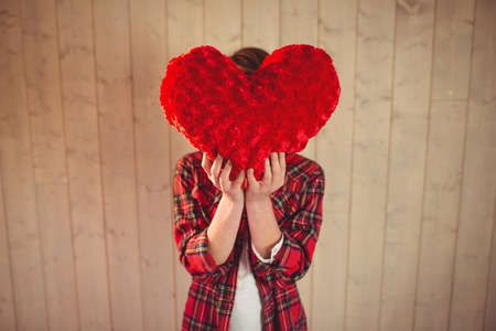 Pretty young woman hiding her face behind heart pillow on wooden planks backgroundの写真素材