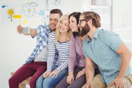Smiling business people taking self portrait on smartphone while sitting on desk in officeの写真素材