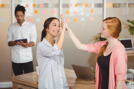 Happy businesswomen giving high five in office with male colleague working in background