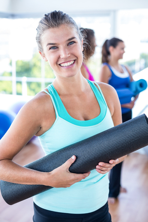 Portrait of beautiful woman smiling while holding exercise mat in fitness studioの写真素材