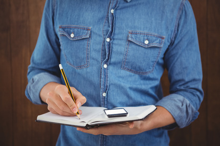 Mid section of hipster with mobile phone writing on book against wooden wall