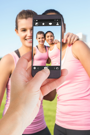 Female hand holding a smartphone against two smiling women wearing pink for breast cancerの写真素材
