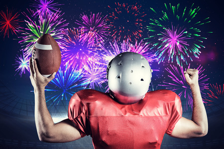 Rear view of American football player cheering while holding ball against fireworks exploding over football stadiumの写真素材