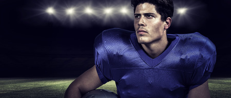 American football player holding helmet while looking away against rugby stadiumの写真素材
