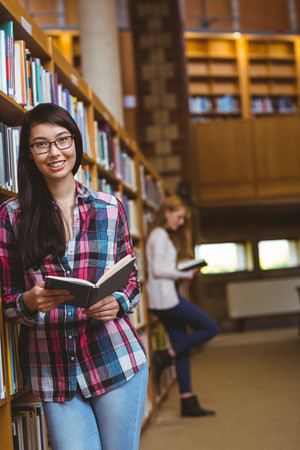 Smiling student leaning against bookshelves reading book at the universityの写真素材