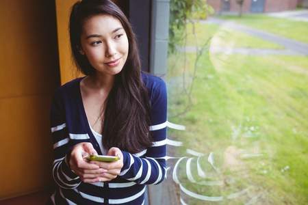 Smiling student with smartphone and looking through the window at the universityの写真素材