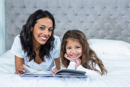 Happy mother and daughter reading a book on the bed at homeの写真素材