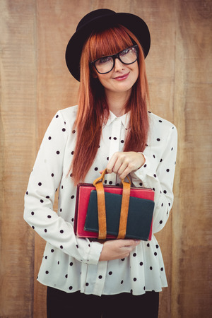 Smiling hipster woman holding book belt against wooden backgroundの写真素材