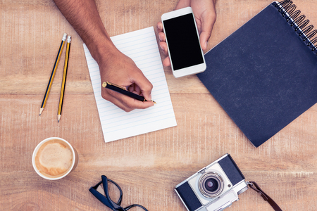 Overhead view of businessman writing on notepad while holding smart phone by camera and eyeglasses on tableの写真素材
