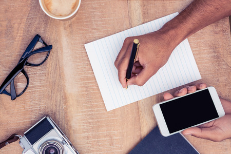 Cropped image of businessman writing on notepad while holding smart phone by camera and eyeglasses on tableの写真素材