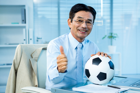 Asian businessman holding soccer ball with thumps up in officeの写真素材