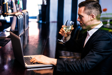Businessman getting a glass of wine and using laptop in a barの写真素材