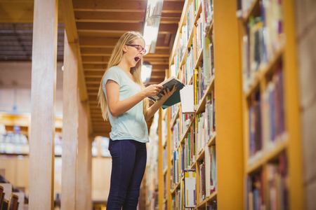 Blonde student reading book next to bookshelf in libraryの写真素材