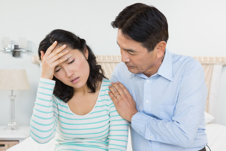 Woman with head ache sitting on bed with husband next to herの写真素材