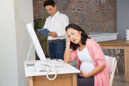 Suffering pregnant woman sitting at desk at home while husband is using tabletの写真素材