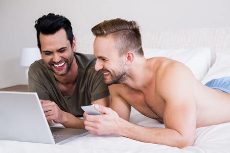 Smiling gay couple using laptop and phone on bedの写真素材