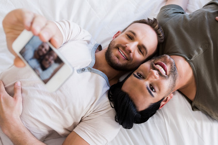 Happy gay couple taking selfie on bed at home