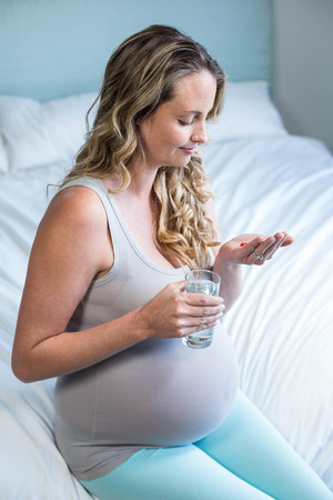 Pregnant woman taking a pill with water in her bedroomの写真素材