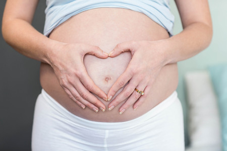 Pregnant woman doing a heart with her hand on her belly in her bedroom