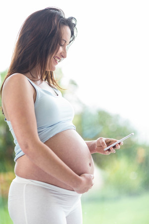 Pregnant woman touching her belly and texting in front of a windowの写真素材