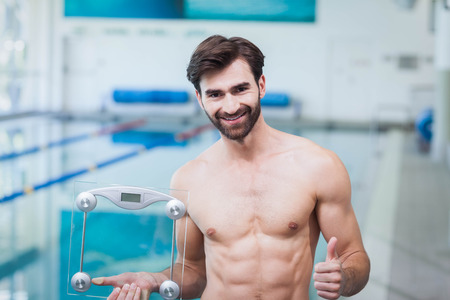 Fit man holding a weighting scale with thumbs up at the poolの写真素材