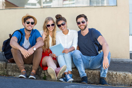 Hip friends holding tablet and laughing while sitting on sidewalk