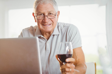 Focused senior man using laptop and drinking wine at homeの写真素材