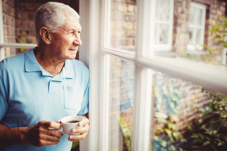 Senior man holding cup and looking out of the window at homeの写真素材