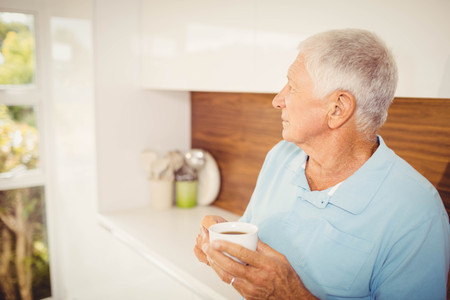 Senior man holding cup and looking away at homeの写真素材