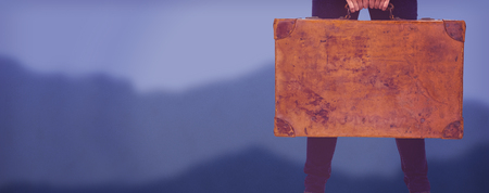 Mid section of a smiling hipster woman holding suitcase against blurred mountainsの写真素材