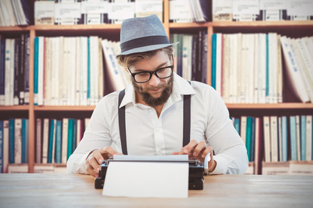 Hipster wearing eye glasses and hat working on typewriter against female student against bookshelf reading a book on the library floorの写真素材