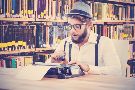 Hipster holding smoking pipe while working on typewriter against close up of a bookshelf
