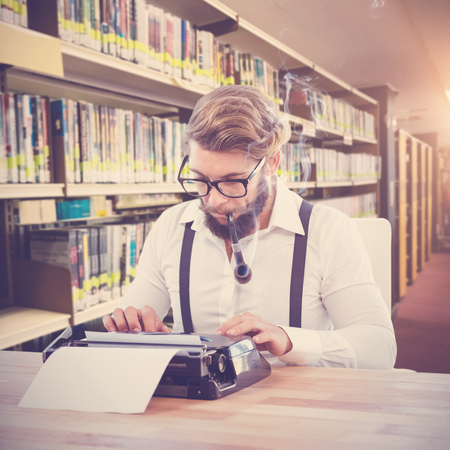 Hipster smoking pipe while working on typewriter against rows of bookshelves in the libraryの写真素材