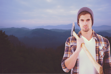 Portrait of hipster holding axe on shoulder against trees and mountain range against cloudscapeの写真素材